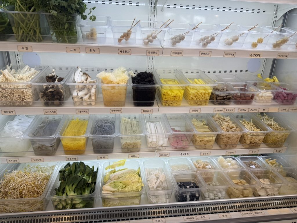 The ingredient bar at a malatang shop: refrigerated bins of mushrooms, noodles, fish cakes, greens and skewers, each labelled in Korean.