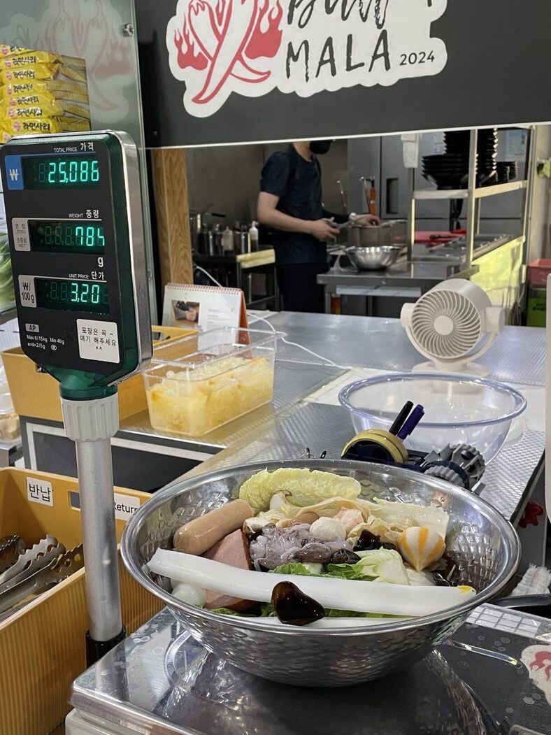 A steel bowl of chosen malatang ingredients on the shop's scale. The display reads 25,086 won for 784 grams.