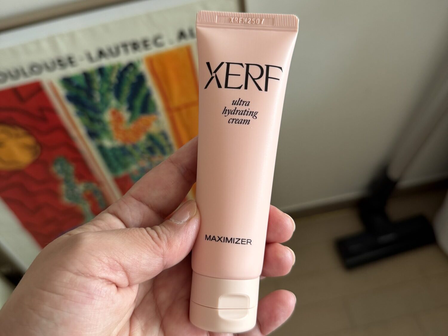 A hand holding a pink tube of XERF ultra hydrating cream, a poster on the wall behind.