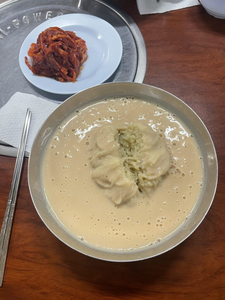 A steel bowl of kongguksu: wheat noodles under a thick, pale soybean broth, with a plate of kimchi beside it.