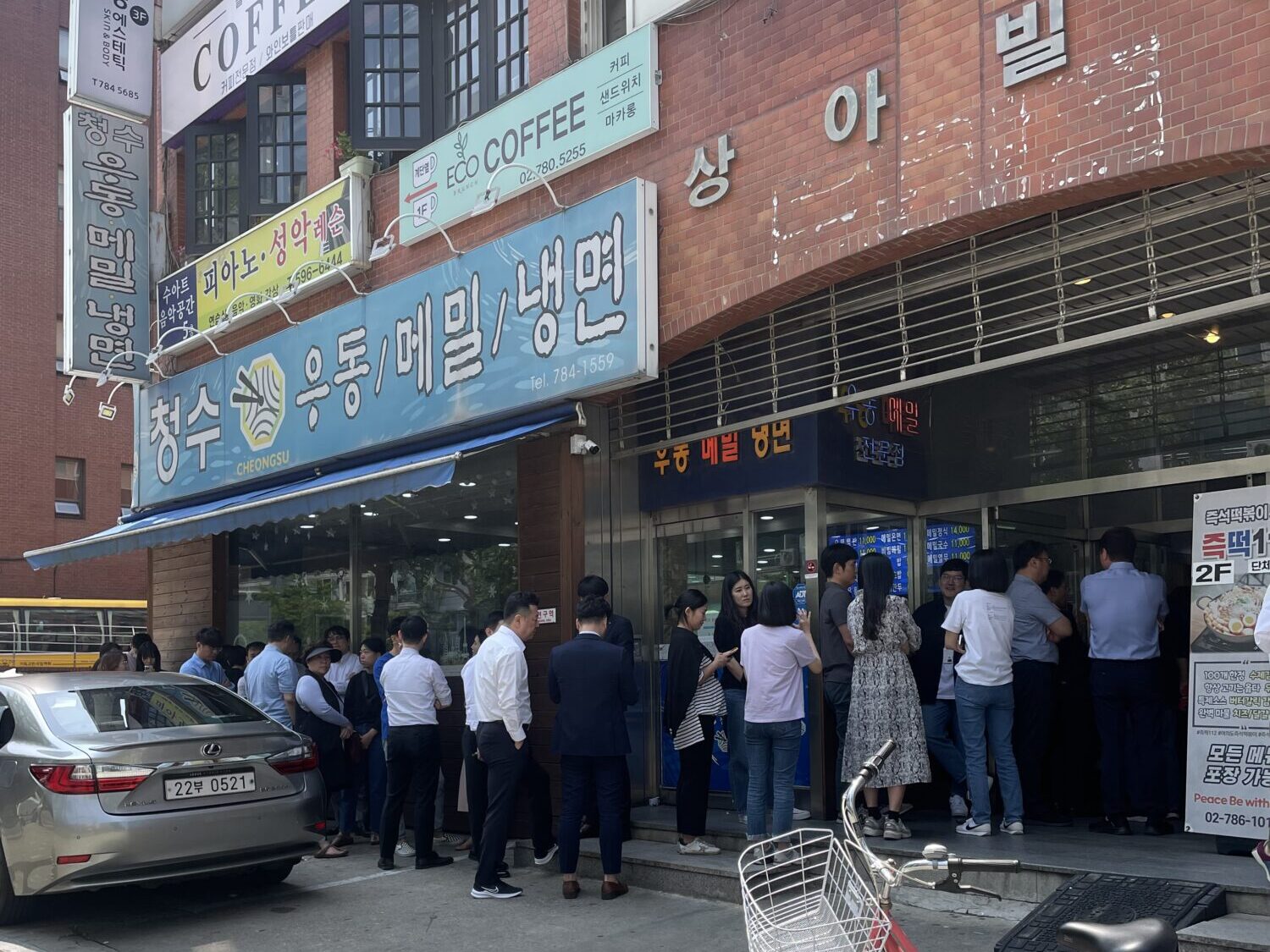 A lunchtime queue on the pavement outside Cheongsu, under a sign reading udon, memil and naengmyeon.