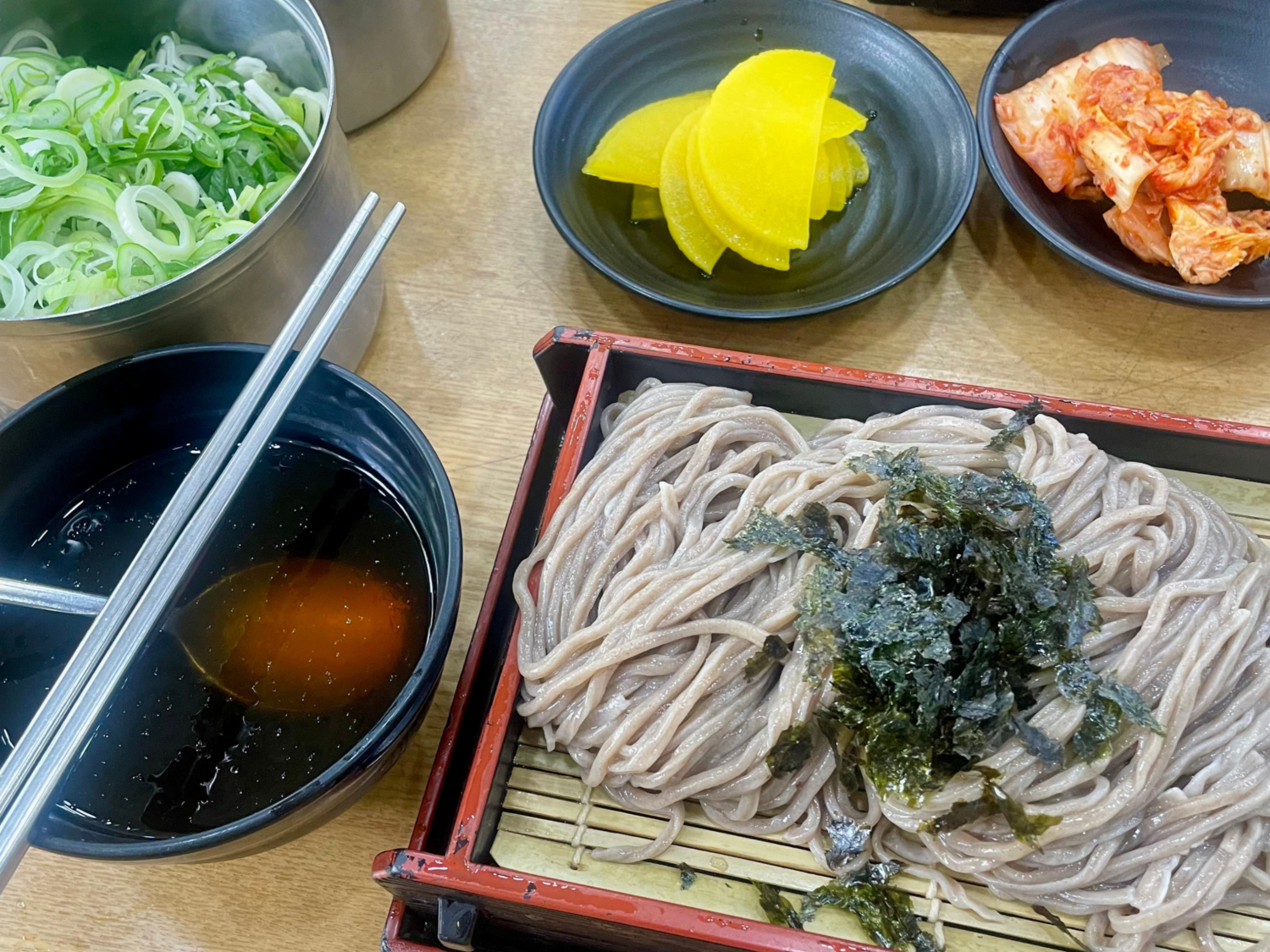 The momil table closer in: chopsticks resting on the bowl of dipping broth, scallion, pickled radish and kimchi beside the tray.