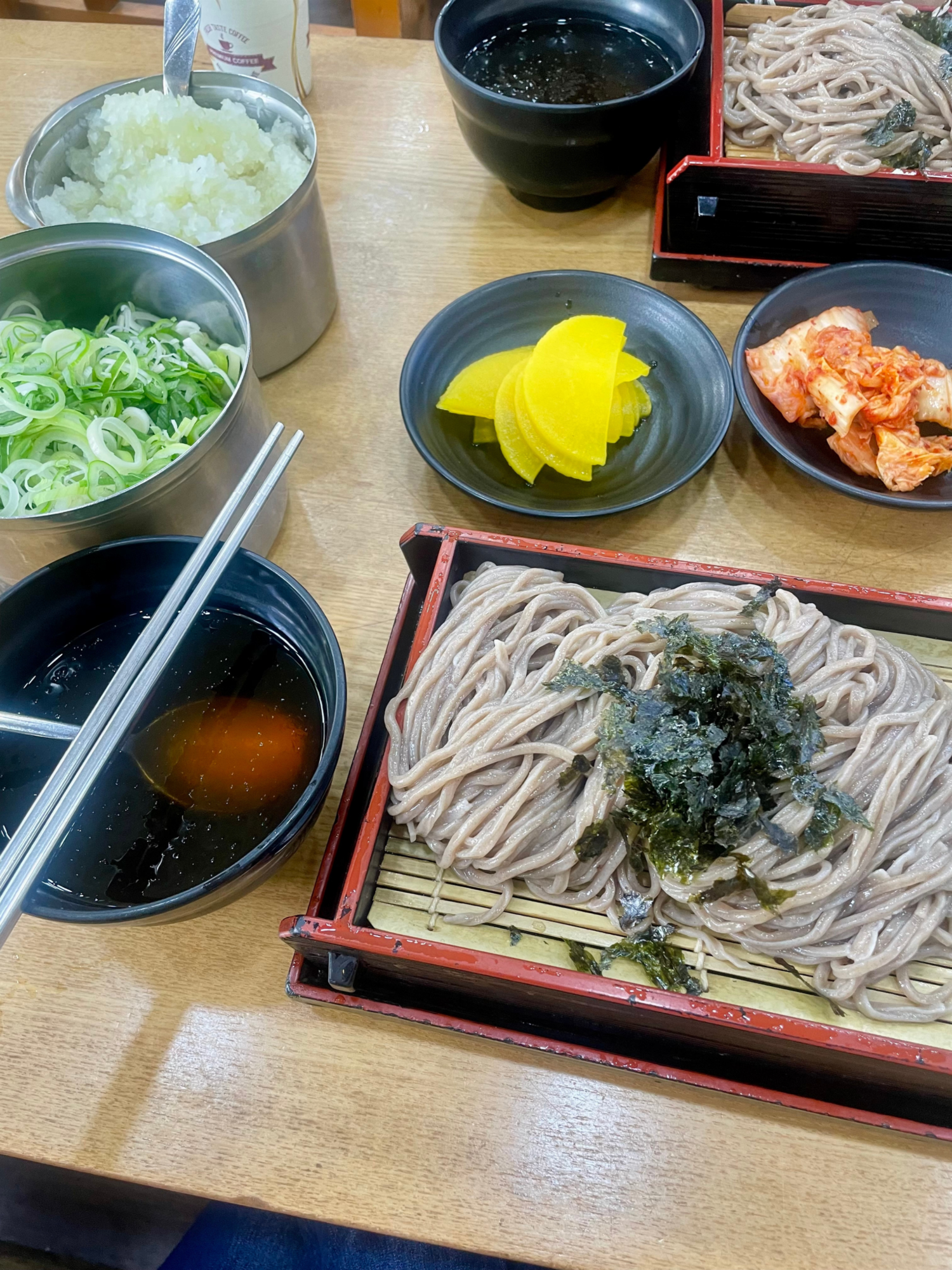 A momil set from above: buckwheat noodles on a bamboo tray, dipping broth, chopped scallion, grated radish, pickled radish and kimchi.
