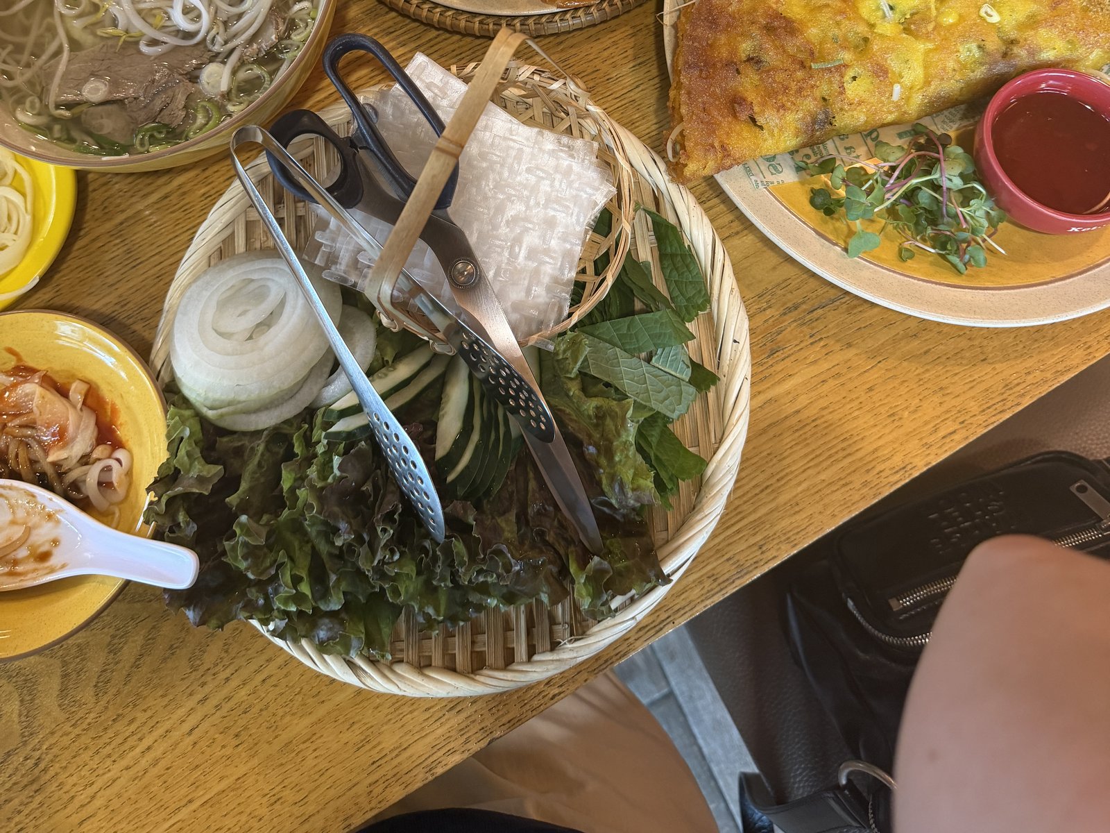 The full table: the pho bowl, the plate of nem, the banh xeo, and a bamboo basket of lettuce, perilla, cucumber and onion with kitchen scissors and tongs laid across it.