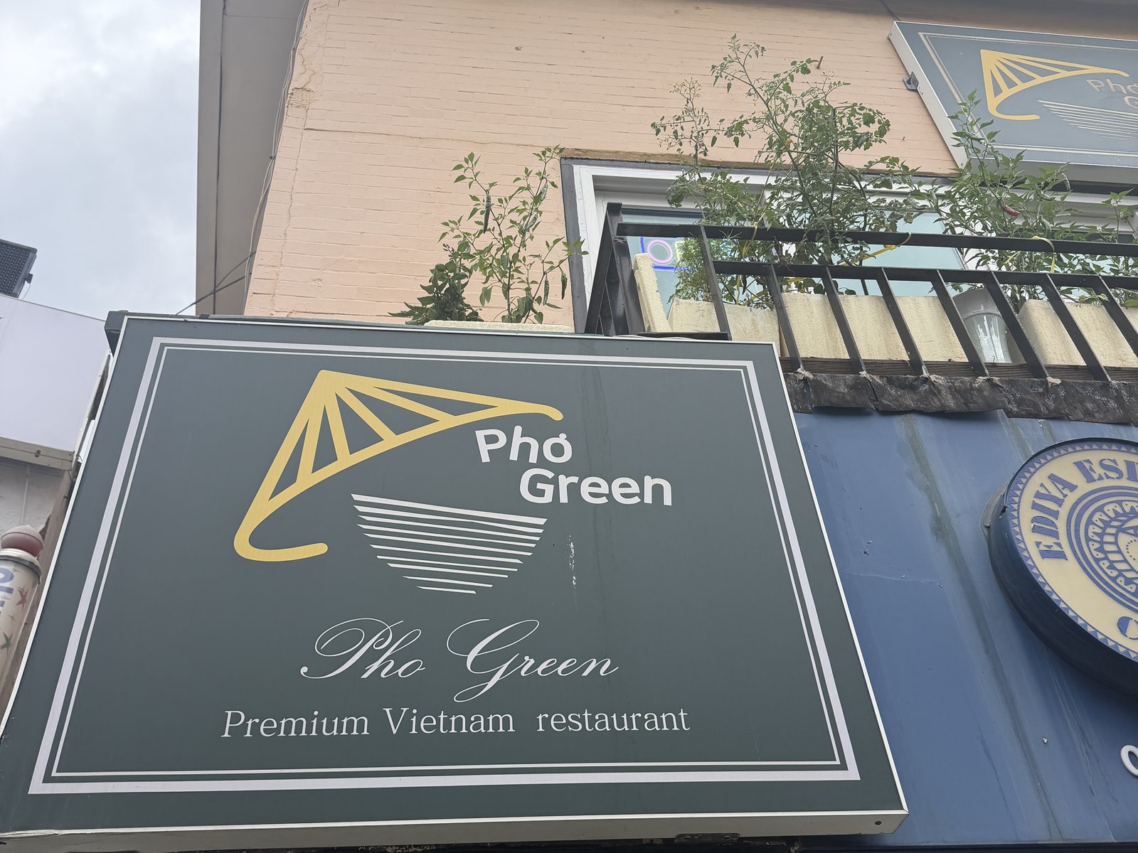 The dark green Pho Green sign on the building front, a yellow conical hat over a noodle bowl, with the Ediya coffee sign next to it.