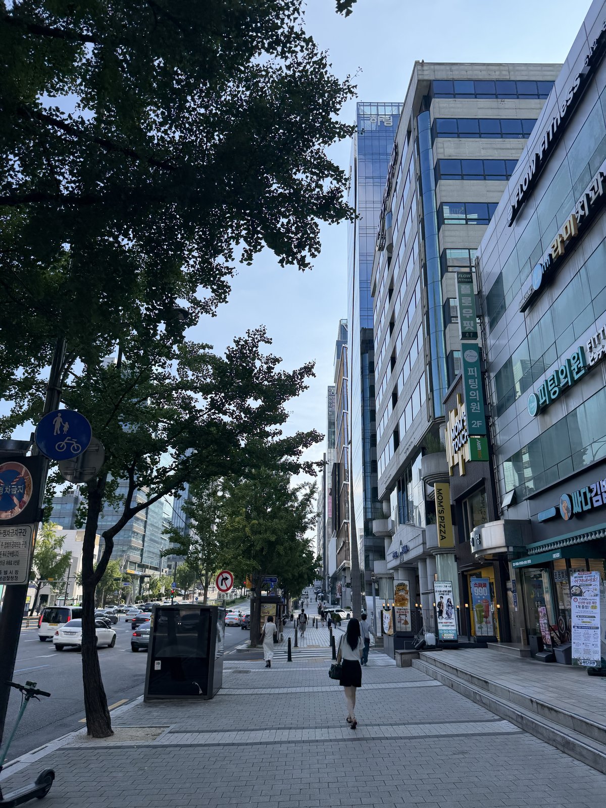 A Seoul office street in the morning, shop signs stacked up the fronts of the buildings and people walking to work.
